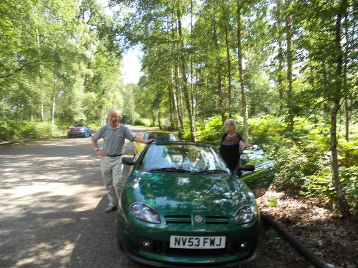 MX5 or MG TF? - Page 1 - MG - PistonHeads - This image shows a man and a woman standing next to a bright green sports car. The car has the make "NISSAN" on the grille and a license plate visible in the bottom-right corner with the letters "NIV53". The vehicle is parked on the side of a road, which is surrounded by trees, indicating a rural or semi-rural setting. The sky appears to be clear and it's a sunny day. There are other cars in the distance, giving a sense of scale to the scene. The man and woman seem to be posing for a photo, with a light and joyful atmosphere.