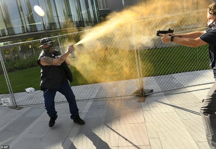 The image depicts a dynamic scene with two individuals in the foreground. One person is aiming and shooting a water gun at another, who is spraying a golden liquid from an unseen container. Both appear to be participating in some sort of game or activity. In the background, there's a fence and what seems to be a crowd of people watching the event. The atmosphere suggests a public space, possibly during a special event or festival.