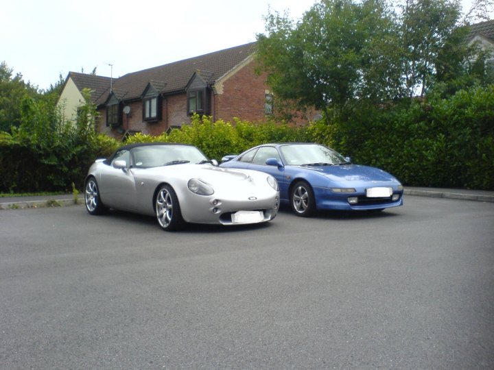 She's a write off. - Page 2 - Jap Chat - PistonHeads - The image features two classic sports cars parked in front of a house with brick walls and lush green shrubbery. The white car, parked on the left, is an Audi R8 and has a sleek, sporty design with five-pointed star emblem. Next to it, a blue car, possibly a longer-wheelbase version of the Audi R8, is uniformly colored with a similar design, side by side on a street with no people visible, suggesting a quiet, residential area. The photo appears to be taken during the day with ample light illuminating the cars and the surrounding environment.