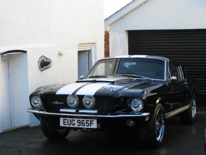 A car parked on the side of the road - Pistonheads - The image features a black and white Ford Mustang car parked in a garage or driveway of a white building. The Mustang is a classic model, known for its distinctive design and powerful performance. It has a sleek body with a striped hood that adds a dynamic touch. The car is positioned at a three-quarter angle, giving a clear view of its front and left side. The license plate is visible, reading "EUG 965F". The setting appears to be a suburban area, with the building waiting to be painted.