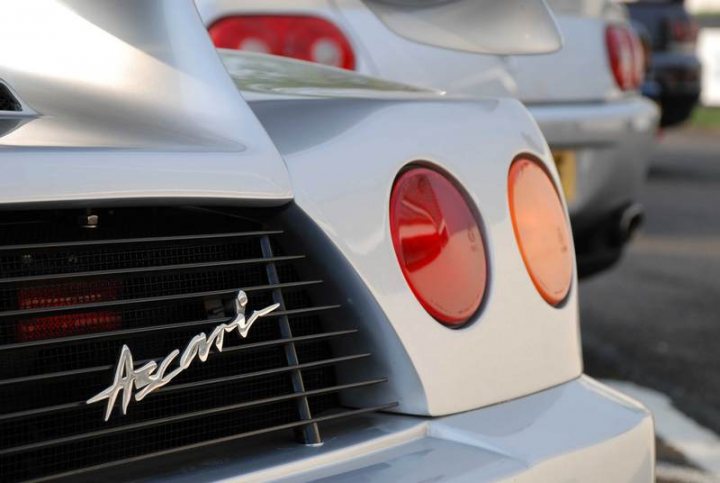 Pistonheads Supercar Sunday - The image depicts a close-up view of a white luxury car with red brake lights. The tail lights are prominently featuring, with the left one a slightly different shade of red than the one on the right. Below the brake lights, there's a silver emblem that reads "Ascari," indicating the make of the vehicle. The image is taken from a rear angle, showcasing the design and features of the car, including the exhaust system directly underneath the tail lights. The backdrop is a blurred scene of a city street.