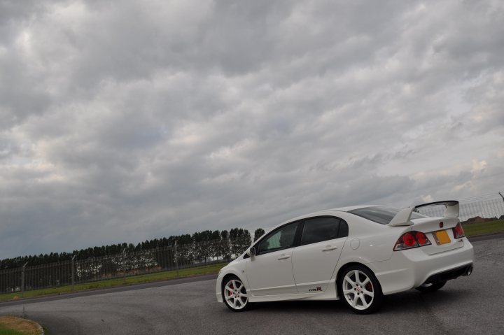 Import Pistonheads Type - The image features a white coupe taking a left-hand bend on a road. The car is captured in motion, and the angle of the photograph gives a dynamic sense of speed. The sky above the car is overcast with gray, moody clouds, and the road ahead appears to be in a rural or semi-rural area based on the presence of trees lining the road and a fence and buildings to the left. The car has a prominent rear spoiler, hinting at its sporty nature.