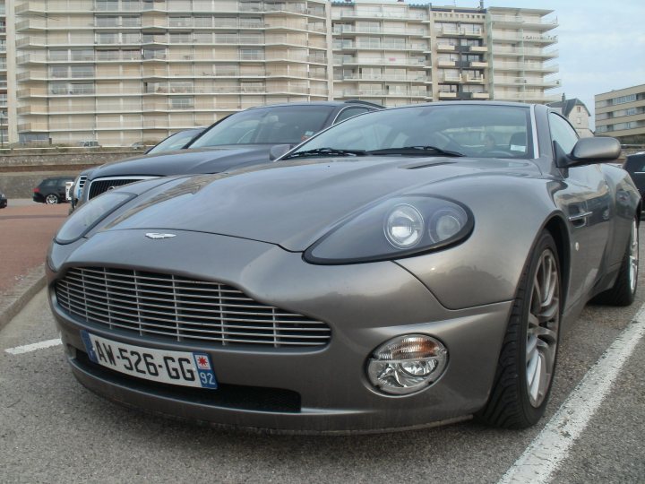 France Spotted Thread! - Page 1 - France - PistonHeads - The image captures a scene in a parking lot where two luxury cars are parked. The car in focus is a gray sports vehicle with a sleek design, prominently featuring a badge that appears to be a logo. Adjacent to it is a large, parked SUV that contrasts the sports car's design with its utilitarian finish. The license plates on both cars are visible, although the specific details are not clear due to angle and proportion. In the background, there are apartment buildings, suggesting an urban setting, and other cars can be seen in the parking lot. The sky above is overcast, lending a cool tone to the overall scene.