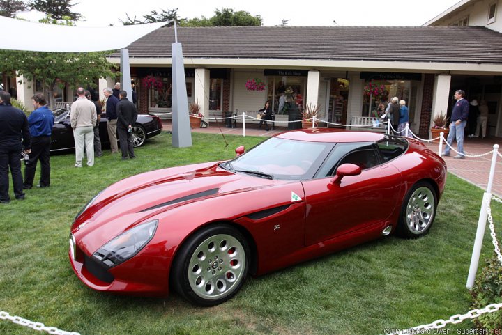 Alfa TZ3 Stradale - Page 1 - Vipers - PistonHeads - The image depicts a dynamic outdoor scene, featuring a striking red sports car as the central subject. The car is parked on a manicured lawn, facing the camera, highlighting its sleek design. Surrounding the vehicle are several people, all attentively observing the car. The setting includes a building with a white roof in the background, suggesting a formal event or showroom. The chrome wheels of the car gleam under the light, indicating a shiny and well-maintained vehicle, likely a high-end model.