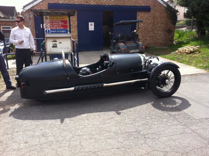 3 wheeler - Page 1 - Morgan - PistonHeads - The image captures an outdoor scene featuring an antique hot rod car. The car, with its classic design and chrome finishing, is parked on a road in front of an old brick building. A man is standing nearby, dressed in a white shirt and sunglasses, contributing to the nostalgic atmosphere. Further enhancing the vintage feel is the presence of a gas pump next to the car, and a sign on the building displaying "Petrol" and "Station Mastery" with "No Stunting, Finals". The overall setting evokes a sense of history and the charm of a bygone era.