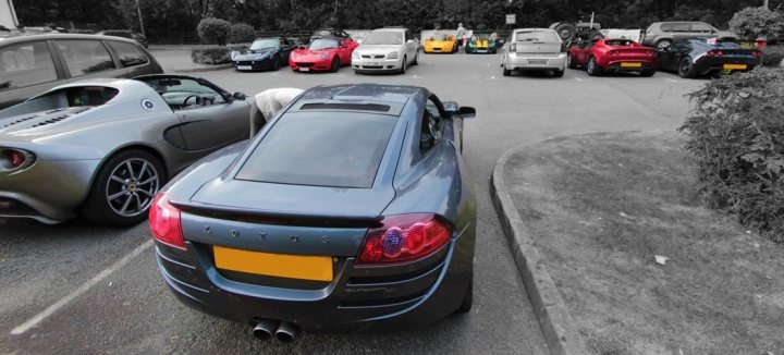 I enjoyed your Lotus North Wales run this evening :) - Page 1 - General Lotus Stuff - PistonHeads - The image is a black and white photograph of a parking lot filled with a variety of cars. In the foreground, there is a prominent sports car with its back facing the viewer, its tail lights illuminated and its rear licence plate clearly visible. The rest of the lot is occupied by several other cars parked in rows. There are trees alongside the parked vehicles, and the view of the cars is framed by greenery in the background. The presence of a rearview mirror allows for a sense of directionality within the image. The photograph conveys a quiet moment in an urban setting, with the focus on the details of the sports car.