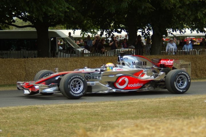 FOS good photography vantage points.. - Page 1 - Goodwood Events - PistonHeads - In the heart of a vibrant motorsport event, a Vodacom-sponsored Formula One car is the star of the show. This sleek, angular car is painted in a striking livery of red, white, and black, its silver and black body gleaming under the stadium lights. The car is moving, seemingly in high gear, adding a sense of urgency and excitement to the scene. The stadium itself is filled with spectators standing in the grass, their attention riveted on the speeding car. The atmosphere is one of thrill and anticipation, as the machine rips through the circuit under the watchful eyes of its drivers and mechanics.