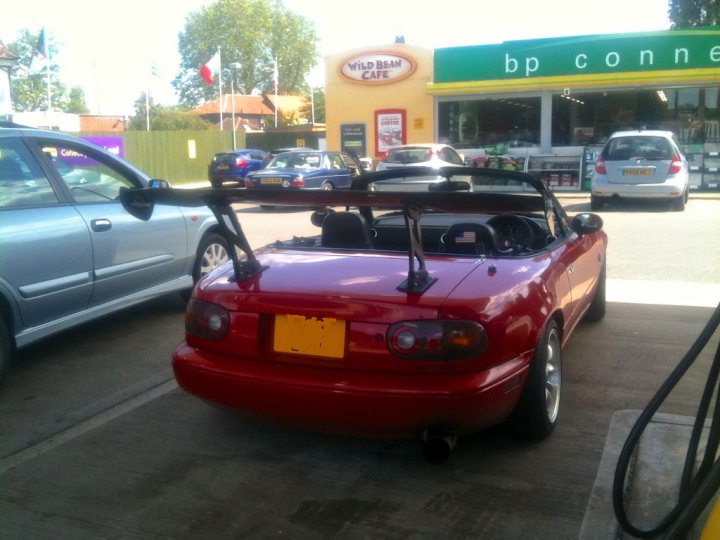 A disturbing sight - Page 1 - Mazda MX5/Eunos/Miata - PistonHeads - The image showcases a vibrant red sports car parked in a gas station parking lot. Its top is down, revealing its interior to be predominantly black fabric, providing a stark contrast to the vehicle's shiny exterior. In the background, there's a gas pump on the right side of the image and a BP convenience store on the left. Other vehicles, including cars and a truck, are scattered in the background, indicating a busy environment.