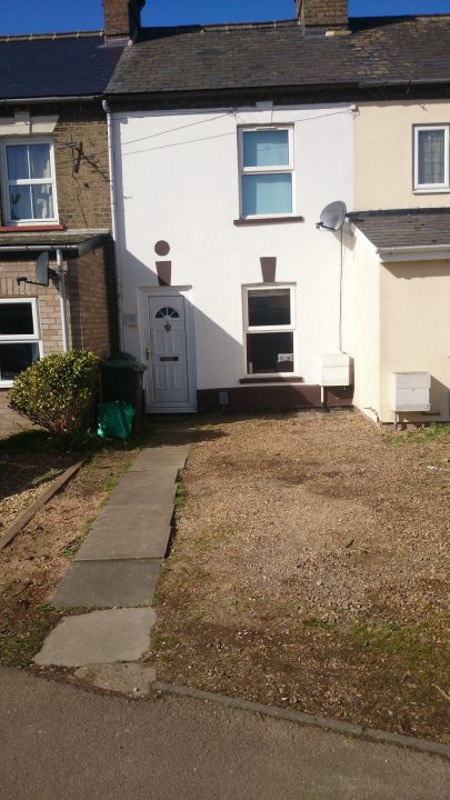 How much to dig out my driveway? - Page 1 - Homes, Gardens and DIY - PistonHeads - The image shows a white house with a brown roof. The house features a white door, which is the central focus. The door is complemented by a green inverted T shaped planter on either side of it. A pathway leads up to the door with a darker section signifying a step. The ground around the house is covered in gravel, and there is a visible satellite dish on a sloping roof above the house. The sky is clear, and the overall setting suggests a residential area.