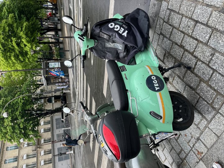 Pistonheads - The image shows a scene from what appears to be an urban setting, specifically on the side of a road. There's a parked scooter with its seat covered by a black item and a bag attached to it, indicating that it might be used for transportation. Adjacent to the scooter is a motorcycle, which is also parked and has a white cover over its seat. The background features elements of city infrastructure, including street lamps and a building with storefronts. There's also a tree in the middle of the road, suggesting it might be a pedestrian area. Overall, the image depicts a common urban scene where various modes of transportation are parked on the side of the road.