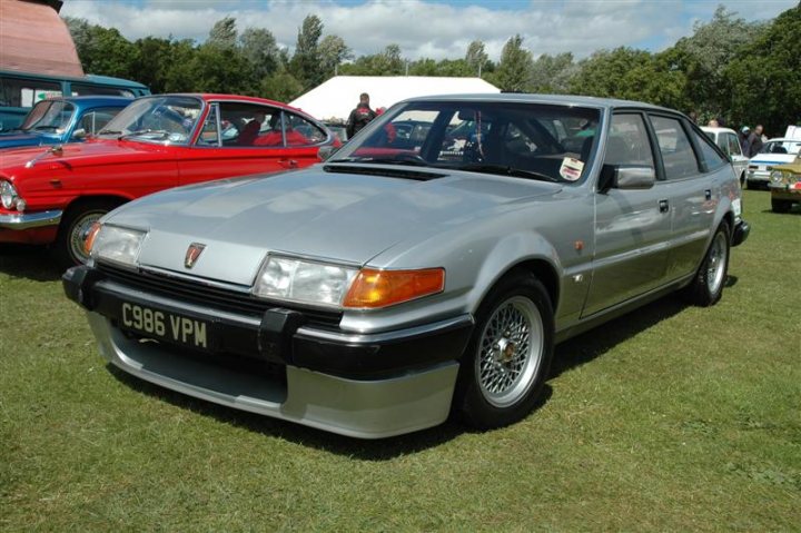 Fins and Chrome - Page 1 - North East - PistonHeads - The image shows a grey car parked on grass, surrounded by various other cars, including a red sports car behind it. The grey car in the forefront appears to be a vintage or classic model, given its distinct design with a prominent grille and large headlights. The weather appears fair with a blue sky, indicating that the photo was taken during the day in good lighting conditions. The setting suggests this might be an outdoor car show or exhibition event. There are a few people visible in the background, possibly spectators or participants, but the focus of the image is clearly on the vintage vehicles.