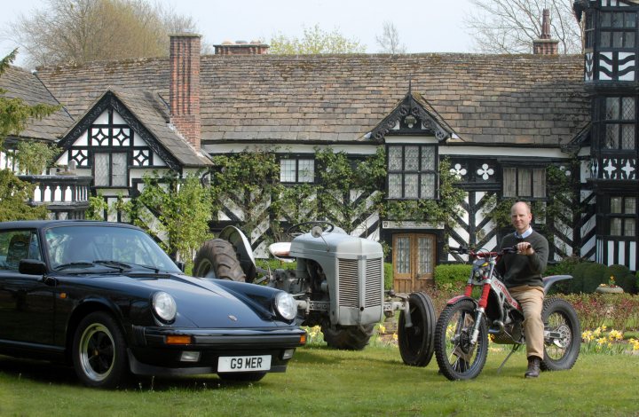 Gawsworth Pistonheads - In an immaculately maintained setting with vibrant colors and lush greenery, a country house with intricate black-and-white beams stands majestically. In front of the house, two vintage vehicles are parked on a green lawn, one a black car with a white license plate, and the other a red motorcycle, adding a nostalgic touch to the scene. A bald man, donning a suit and holding a brochure, stands next to the motorcycle, possibly posing for a photo, in the company of these classic vehicles and the beautifully designed house.