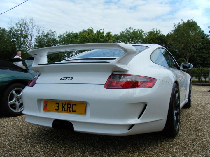 Breakfast Meeting, The Hare, Roxwell 4th June - Page 1 - Kent & Essex - PistonHeads - The image features a white Porsche sports car parked on a gravel driveway. The car's license plate reads "3 KRC", which could indicate the model or registration details. The Porsche has a distinct black rear spoiler, enhancing its sporty design. The vehicle is positioned at an angle that showcases its rear side, emphasizing its aerodynamic elements. In the background, there's a contrasting green car, providing a complete scene of the driveway where the Porsche is parked.