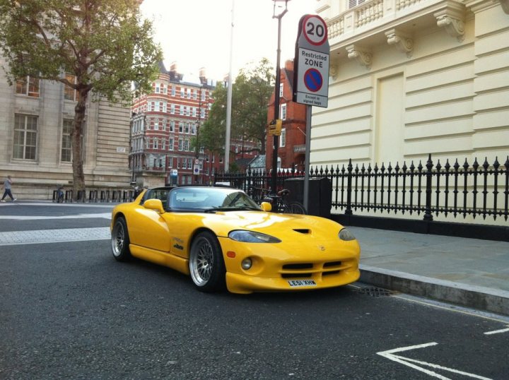 A car is parked on the side of the street - Pistonheads - The image shows a bright yellow convertible sports car parked on the side of a street next to a paved area. The car is yellow with black accents and wheels. The street scene includes a pedestrian on the sidewalk, a red and white speed limit sign, and a black wrought-iron fence in the foreground. The buildings in the background appear to be tall and older, suggesting an urban environment, possibly in Europe.