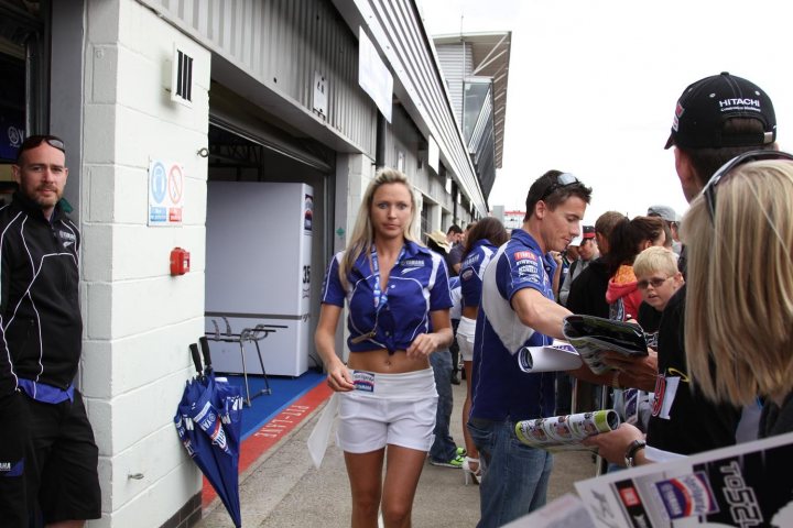 - Page 307 - General Gassing - PistonHeads - This image captures a dynamic scene inside a building. At the forefront, a blonde woman in a blue and white uniform is walking towards the camera. Behind her, a man and woman in matching uniforms are interacting, while a crowd of people, some of whom are holding umbrellas, watches the action. A few items, including a can, a black and white hat, and a chair, can also be seen in the image. The environment suggests an event or gathering is taking place.