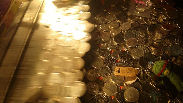 The official Nokia EOS thread - Page 3 - Computers, Gadgets & Stuff - PistonHeads - The image is slightly blurry, with coinage spread out on what appears to be a hard surface, possibly a table or the floor. In the center of the coin pile, there is a small brown cardboard box resting on the coins. A colorful, yarn-like creature, which could be a green toy from a game or a fabric item, is intertwined among the coins on the right side of the image. The display is illuminated by light, casting a reflective sheen on the coins and adding to the metallic look of the scene. The overall impression is of a playful or whimsical setting with a focus on the contrast between the small, obscure, and vibrant object among the orderly arrangement of the currency.