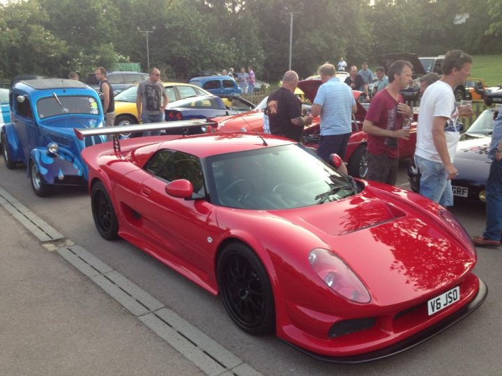 South Bucks Car meet Tuesday 13th August - Page 1 - Thames Valley & Surrey - PistonHeads - The image showcases an outdoor auto show where several cars are on display. The prominent car in the foreground is a vibrant, polished red sports car, featuring a sleek design with black rims and a zero-emission license plate. The car is parked parallel to the road and has a distinctive front grille and rear spoiler, both suggesting high performance capabilities. In the background, people are walking around, interacting with other classic cars. The event appears to be well-attended, with other vehicles parked behind the red sports car, indicating the variety and scale of the auto show.