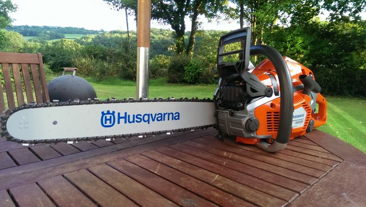 Small chainsaw - Husqvarna, Stihl or Makita ? - Page 2 - Homes, Gardens and DIY - PistonHeads - The image depicts an orange and black chainsaw lying idle on a wooden table outdoors. The table is placed on a patch of grass with green woods in the background. The chainsaw's handle is pointing towards the top right corner of the photo. Upon the chainsaw, a black and white bumper sticker is visible bearing the name "HUSQVARNA," a Swedish company well-known for producing high-quality chainsaws. The focus of the image is on the chainsaw, drawing immediate attention to it.