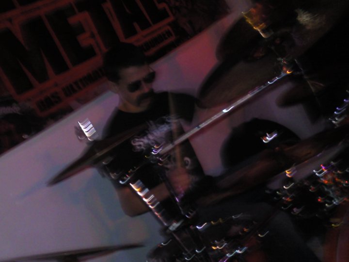 Gig Pistonheads Lets - The image is a highly blurred, motion-blurred photograph of a person playing a drum set. The lighting and vivid colors give the image an energetic feel. A person, wearing sunglasses, is focused on the drums, suggesting a live performance. The background is relatively out of focus, emphasizing the action of the drummer. The motion blur effect creates a sense of dynamism in the image.