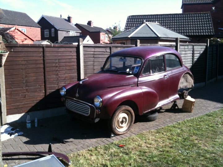 1971 Morris Minor 1000 Resto thread (Maybe) - Page 1 - Classic Cars and Yesterday's Heroes - PistonHeads - In the image, a burgundy classic car is the centerpiece, parked on a grassy area with a wooden fence in its immediate surroundings. The car, with its vintage appeal, features a luggage rack on its rear, adding to its distinctive charm. Beyond the fence, traditional houses can be seen, suggesting a suburban setting for this scene. The backdrop of the sky, visible through the topmost part of the image, adds a tranquil touch to the scene.