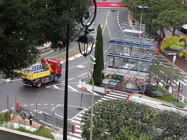 A truck with a trailer attached to the back of it - Pistonheads - The image portrays an urban scene featuring a street with crosswalks and traffic signs. A large, orange and yellow construction truck is in the middle of the road, directed by traffic lights. To the right of the truck, there is a tall, temporary scaffolding structure, standing at an intersection. A couple of traffic lights are visible, guiding the traffic. Around the area, there are plants of various sizes, adding to the cityscape. There are also several people visible on the sidewalks, indicative of typical urban life.