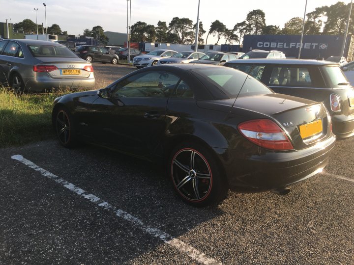 R171 SLK200 6 Speed Man 1.8 L M271 - Page 1 - Readers' Cars - PistonHeads UK - The image shows a black sports car parked in a parking lot. The vehicle has distinctive features such as large rims and a sleek design. It appears to be a two-door coupe, suggesting it is designed for driving enthusiasts or for those who prefer the agility of smaller vehicles. The car is parked next to a marked parking space. In the background, there are other cars in the lot and what looks like a building with the word "Sainsbury" visible on it. The lighting suggests either early morning or late afternoon, as indicated by the soft light cast across the scene.