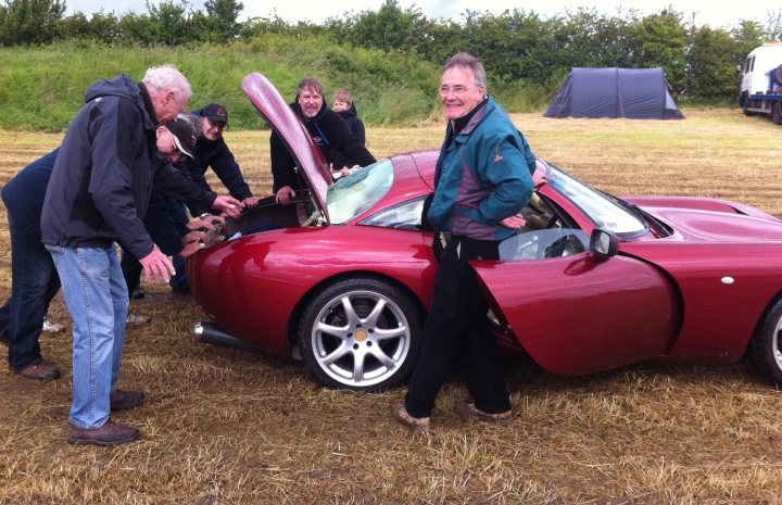 Old Buckingham air show - Page 1 - East Anglia - PistonHeads - In the image, a group of people are inspecting a sleek, red sports car. The car is parked on a grassy area filled with hay. The group appears to be engaged in conversation or discussion, possibly about the car. The car itself is elegant, with a distinctively streamlined design that suggests speed and performance. This image captures a moment of shared interest and engagement among a small group of people and a luxury car.