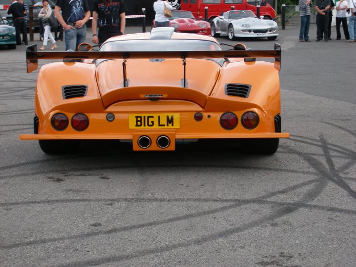 Pistonheads Evo Marcos Restored Ace Appears Cafe - In the image, there is a slick orange sports car parked on a street. The car is a two-door racer with a large rear wing, which is a distinctive feature. The license plate reads "BIG LM." In the background, there are several other cars and people, suggesting this could be a public area or open car show. The ground is marked with tire tracks, possibly from other vehicles that passed by. The car appears to be a racer with a powerful engine, as indicated by the design.