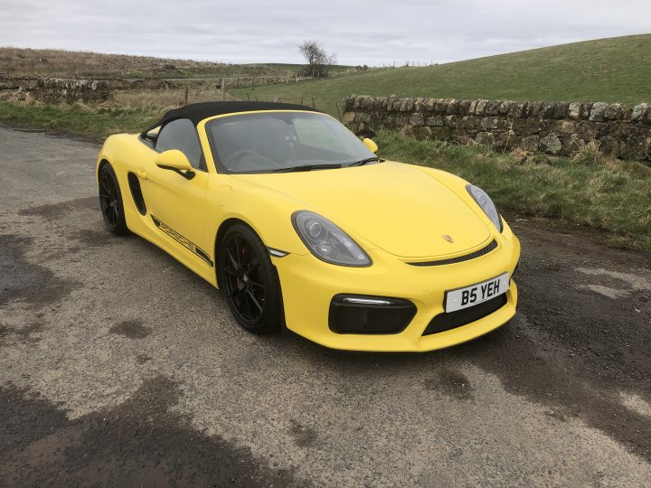 Who else is getting their Summer cars out?  - Page 7 - General Gassing - PistonHeads UK - The image depicts a vibrant yellow sports car parked on a road. The car is a convertible with the top down, showcasing its sleek design. It's positioned near the side of the road, with a grassy area and a stone wall visible in the background. A license plate is prominently displayed at the back of the vehicle, and there is no text or branding visible on the car itself. The setting suggests a rural or semi-rural location, given the sparseness of the surroundings.