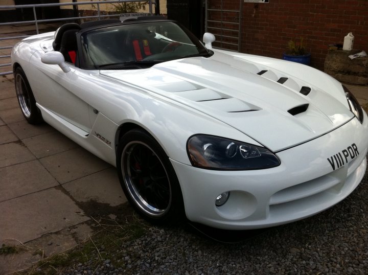 Those side exit exhausts... - Page 1 - Vipers - PistonHeads - The image shows a white sports car parked on a paved surface. The car has a sleek design, characterized by a large, probable wind deflector or air-intake vent on the hood. The front of the car is oriented towards the left side of the image, and it features prominent headlights and a vented hood that gives it a powerful and sporty appearance. The tag "VVIPR" is visible on the grill, and the vehicle is situated in front of a building with a brick wall.