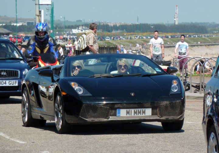 A group of people riding on the back of a motorcycle - Pistonheads - In the image, a group of people are riding down a street on a day that appears to be quite warm and sunny, as suggested by their attire. Leading the group is a dark-colored convertible sports car, with another car closely following behind. The road they're on appears to be in a city or town, as indicated by a mix of pedestrians and other vehicles visible in the background. The sky is clear, and the daylight is bright, suggesting it might be summer.