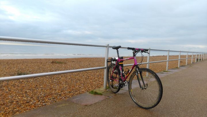 The "Photos From Today's Ride" thread... - Page 1 - Pedal Powered - PistonHeads - The image presents a serene beach scene, dominated by a choice of bicycle parked on the sidewalk near a metal barrier. The bicycle, characterized by its purple frame and matching black tires, stands tall and unattended. The beach in the backdrop features pale, light-colored sand that extends towards the horizon. Above, the sky is overcast, suggesting a possibility of rain. The beach is devoid of any apparent human activity. The overall image evokes a sense of solitude and tranquillity.