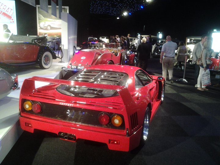 Ferrari F40 Values - Past and Present - Page 9 - Supercar General - PistonHeads - The image shows a vibrant indoor car show. A collection of classic and vintage sports cars dominate the scene, with a striking red Ferrari positioned in the foreground. The cars are parked on carpeted floors, and various attendees can be seen strolling around the exhibition, with some closer to the camera, admiring the displays. The environment is illuminated by spotlights that cast a warm glow over the exhibit, emphasizing the sleek lines of the cars and creating an atmospheric display.