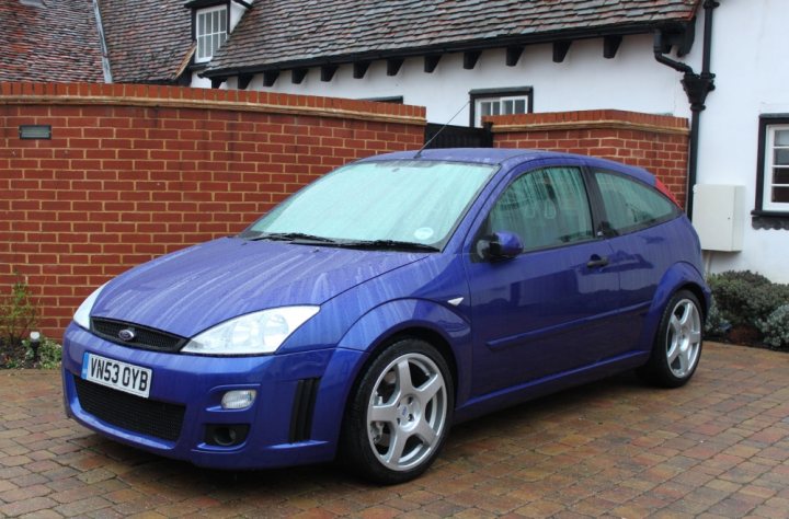 Let's see your fords - Page 21 - Ford - PistonHeads - The image shows a blue Ford Focus parked next to a brick wall. The vehicle appears to be a small hatchback with silver wheels. It's a daytime photo, and the car looks to be in good condition. The license plate is clearly visible on the front of the vehicle. The overall setting suggests a typical residential area.