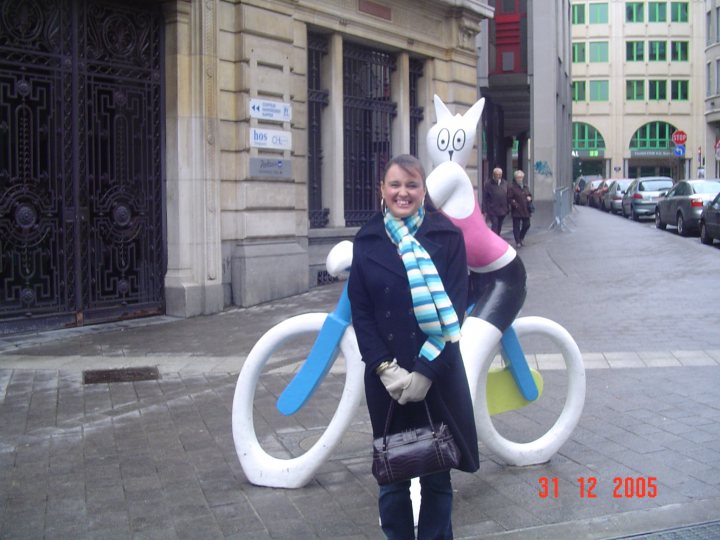 Odd Strange Front Pistonheads Tourist Statues Poses - The image features a woman standing on a sidewalk, smiling at the camera. She is wearing a blue and white striped scarf and has a black purse with her. Behind her, on the wall of a building, is a large sculpture of a cat sitting on a bicycle. The woman is standing in front of a stone building with metal gates, and there are other people walking by and cars parked along the street behind her.