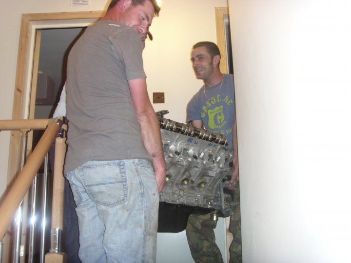 Pistonheads Outriggers - The image shows a domestic scene with two men engaging in different tasks. The man on the left is standing and facing away from the camera, seemingly focused on pulling up his pants or preparing to leave. On the right, a man stands behind him, looking at the camera with a cheerful smile. He is holding a mechanical object that appears to be an engine or large motor, possibly for a car or motorcycle, as suggested by the design and shape. The setting is indoors, as suggested by the walls and floor, and there's a glimpse of a staircase in the background. No text is visible in the image to provide additional context. The overall atmosphere of the image is casual and relaxed.