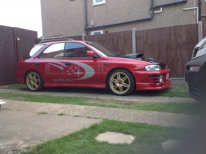 A red car parked in a parking lot - Pistonheads - The image features a vibrant red sports car parked on a gravel driveway next to a fence, with grass on either side. The car has unique customizations, including gold rims, a kit featuring a distinct red and white design adorning its hood, and a black front grill. The vehicle's design suggests it is a model by Subaru. The car holds a dominant position in the scene, with its robust and sleek appearance reflecting the owner's keen interest in style and speed. The setting is ordinary yet vividly complements the car's striking appearance.