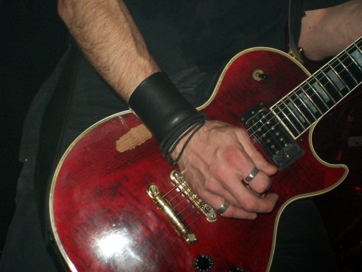 Lets Pistonheads Gig - A person is holding a red electric guitar. They are wearing a black leather wristband, one silver ring, and appear to be wearing black clothing. The focus is on the hand performing with the guitar, specifically on the fingers and the freetails on the green-white-black guitar pickups. The image has a colorful and vivid feel, emphasizing the instrument's details.