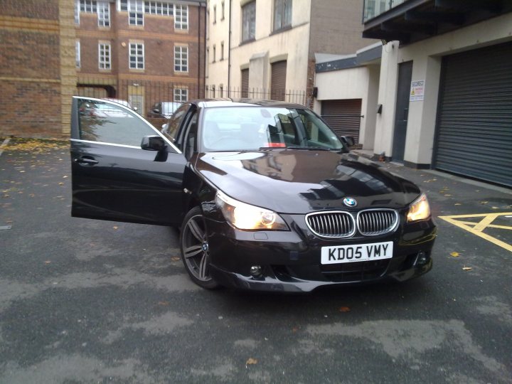 Kevin Pistonheads Bmw - In the image, a black BMW car is parked on a street. The car is positioned in front of an open passenger door. On the hood of the car, there are indications that it is a convertible, with a manually operated convertible top. The car's license plate reads "KD05 VVY". To the right of the car, there is a closed roll-up door, and in the background, there appears to be a brick building. The car is stationary and there are no visible persons in the image.