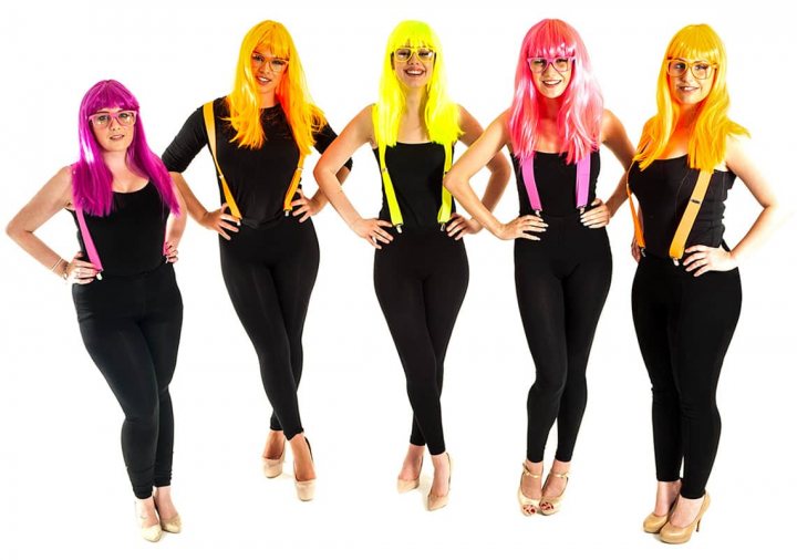 A group of women standing next to each other - The image captures a group of five individuals posing for a photograph. They are all adorned in black jumpsuits with yellow suspenders and sport wildly colored hair in vibrant hues of pink, yellow, green, and purple. The individuals face the camera with wide smiles, creating a cheerful and lively ambiance. The white background accentuates their colorful attire, enhancing the vividness of each character's unique style. The overall image exudes a fun and spirited aesthetic.