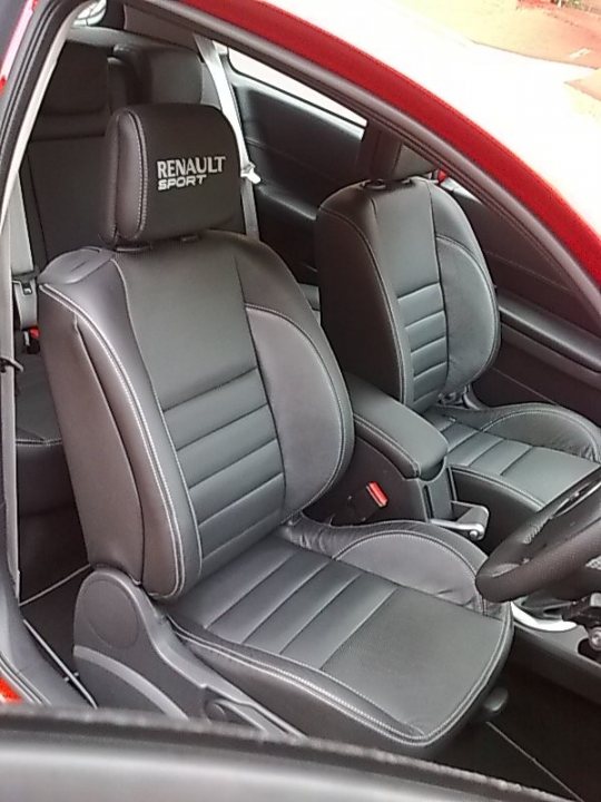 Replacement Pistonheads - This image shows the cabin of a red Renault car. Inside, there is a pair of reclining seats in a dark grey color with leather upholstery. A logo indicates that the vehicle is either new or sellable. The seats are fitted with multiple features such as armrests and headrests, enhancing the car's interior comfort. The steering wheel is also visible, confirming the perspective from the front passenger side.