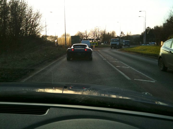 Brand Pistonheads Tesla Spotted - The image shows the interior view from the driver's seat of a car on a highway. The dashboard appears visible, with parts such as the instrument cluster and steering wheel visible. Outside the car, the road is in view, showing other vehicles and landmarks. The car, which bears a blue and white license plate, exhibits the rear end, and the photo's angle gives a glimpse of the surrounding highway environment. The sun somewhat obscures part of the car, suggesting it's taken during the day.