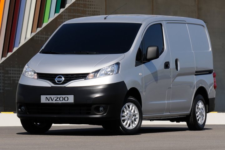 A white van parked in front of a building - Pistonheads - This is a color photograph of a silver Nissan minivan. The vehicle is positioned at a slight angle against a backdrop that features a colorful structure with multiple panels of varying colors. The minivan has a front license plate that reads "NV200" and a side driver's side window location is shouldered by this colorful backdrop. The overall scene suggests a possible introduction or launch event for this vehicle.