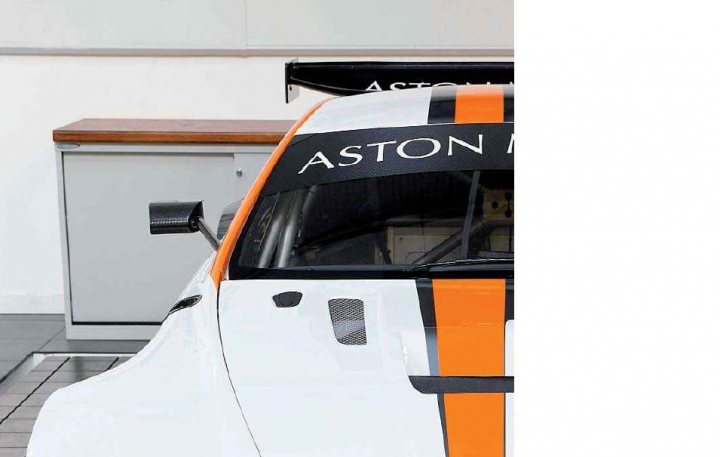 Carbon fibre wing mirrors - GT2 style - Page 1 - Aston Martin - PistonHeads - The image shows the side rear view of a racing car, featuring the Aston Martin logo on the side. It is parked next to a gray filing cabinet or cabinet doors on what appears to be a floor or tiled surface. The car has a large spoiler and a black, orange, and white stripe design elements on its exterior. It seems to be a high-performance vehicle given its sporty aerodynamic details. The background is minimal and focuses attention on the car. The setting suggests a garage or workshop environment.