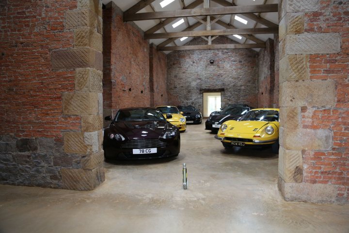 Who has the best Garage on Pistonheads???? - Page 200 - General Gassing - PistonHeads - The image captures a spacious garage with a rustic charm. The garage is filled with a line of shiny, vintage cars, painted in shades of yellow, black, and red. The central room is dominated by stone pillars, while a wooden crossbeam stretches overhead, providing a stark contrast to the building's rough exterior. A single flashlight lies on the cold, concrete floor, adding a sense of mystery. The arrangement of the cars suggests that they are on display, perhaps in preparation for a show or auction. The relative positions of the cars and the flashlight indicate careful planning and presentation.