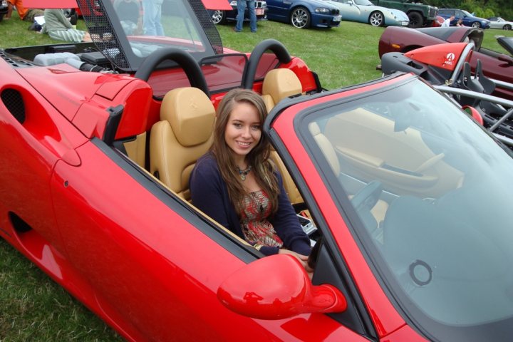 Pistonheads - The image showcases a red sports car with its top down, exhibiting a vibrant red glow in daylight. The view from inside the vehicle provides a unique perspective; we see a young woman in the driver's seat, who is wearing a casual tee shirt and smiling broadly. In the periphery, there are numerous other cars on display, suggesting this scene might be part of a car show or exhibition.