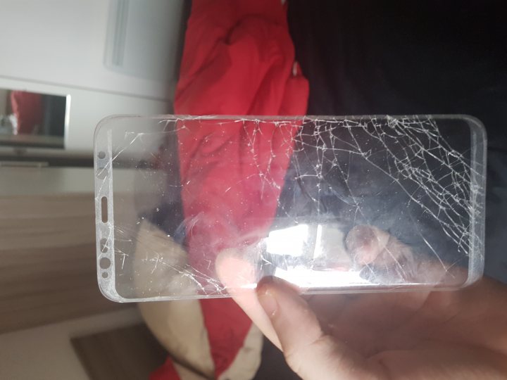 Samsung Galaxy S8 - Page 17 - Computers, Gadgets & Stuff - PistonHeads - The image shows a cracked phone case being held in a person's hand. The case appears to be made of glass or a similar translucent material. The pattern of the cracks radiates from a single point and suggests the phone has been dropped or subjected to force. The person's hand is visible at the bottom of the image, gripping the case. In the background, there's a glimpse of a red garment, possibly a jacket or shirt, indicating an indoor setting.