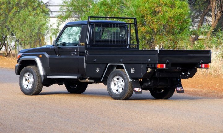 Pistonheads Ute Recommend - The image features a black Toyota Hilux pickup parked on a dirt road. The vehicle exhibits a rugged design with large tires and a raised suspension, suggesting it's designed for off-road use. A distinctive feature of this Hilux model is the addition of a steel cage in the back. Surrounding the truck, there are green trees, indicating an open or woodland area. The overall style of the image seems to be a candid photograph, likely taken in daylight, with the vehicle being the central focus.