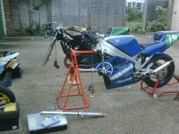 Bikey Tis Pistonheads Weekend - The image features a motorcycle that has been stripped down and is in the process of repair or modification. The motorcycle is the main subject and occupies a significant portion of the frame. It's standing on a wooden stand with an apparatus attached to the front, likely for lifting the front wheel of the motorcycle during maintenance or work. There's a myriad of parts scattered around the immediate vicinity, suggesting ongoing work. The setting appears to be a storage or garage area, with various other items visible in the background.