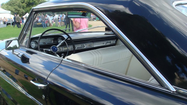 GRRC Open Day - 11 August - Page 1 - Goodwood Events - PistonHeads - The image captures a classic black car, presumably a convertible, parked outdoors. The car's interior is visible through the slightly open window, revealing a white leather seat and a steering wheel. The curved design of the passenger door frame is more detailed than the other doors, suggesting a vintage style. People are standing around the car, creating a sense of life and activity in the scene. This setting appears to be a car show or exhibition, with the vehicle on display.